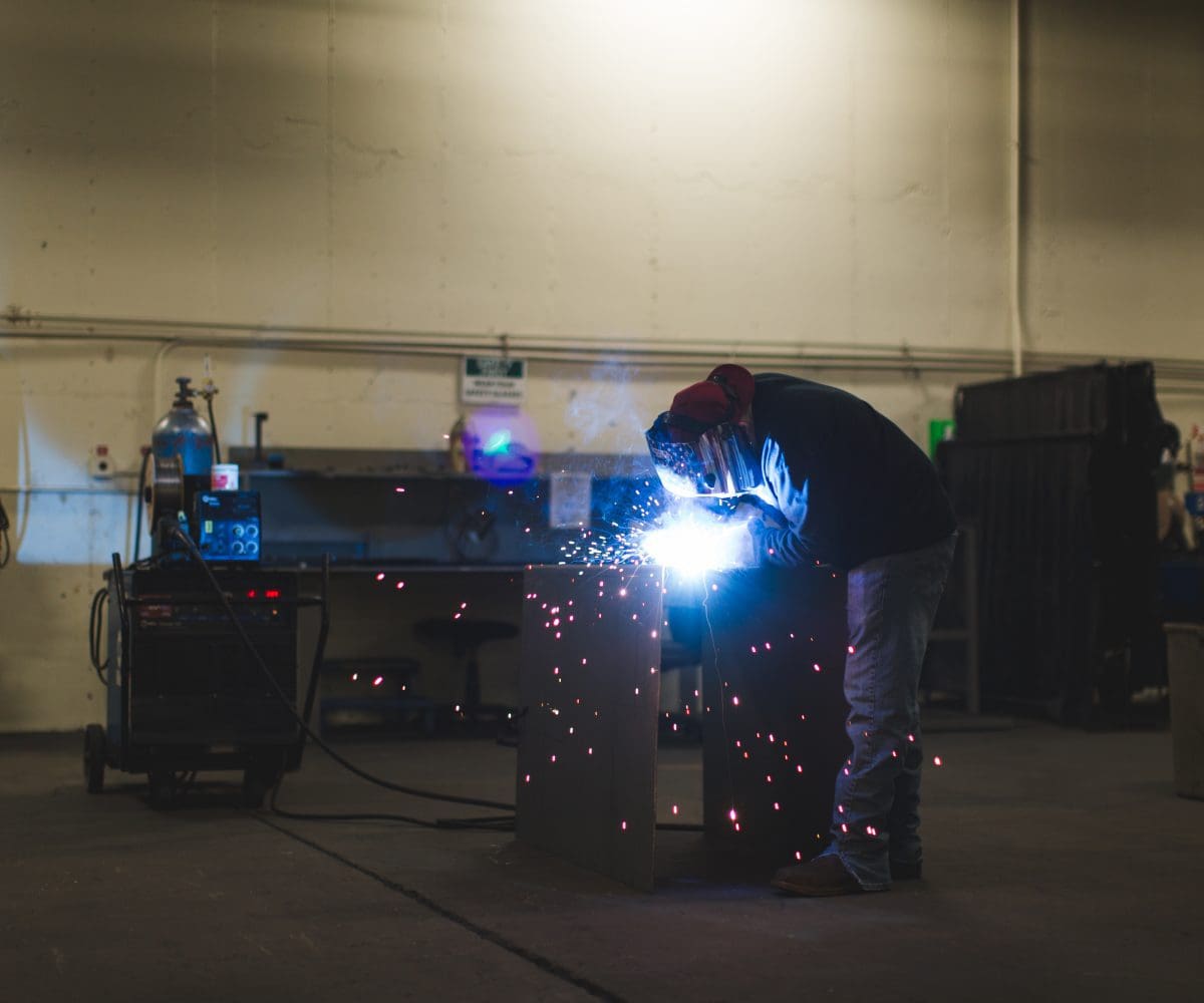 Neff_Press_3_13_shoot_edited-63-1200x1000 A man welding in an industrial setting with lights.